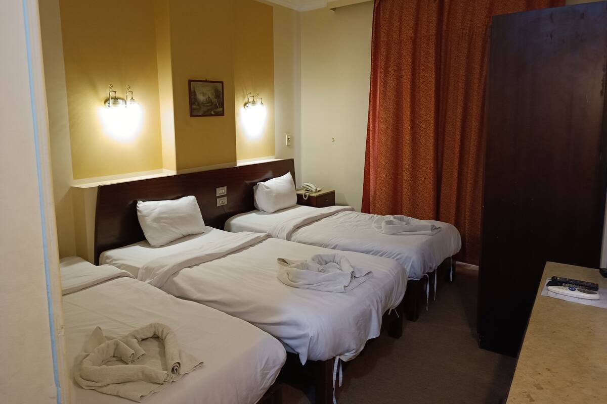 Quarto de hotel com três camas de solteiro, cabeceira de madeira, mesa de cabeceira com telefone e um armário grande de cor escura.