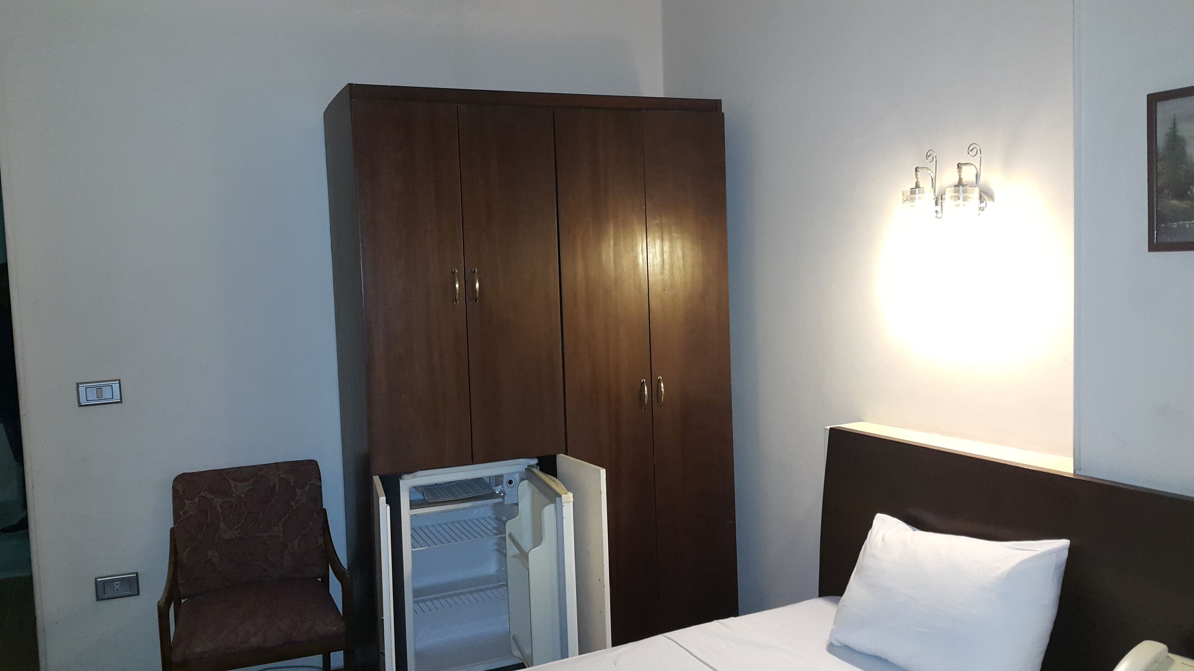 A hotel room with a bed, a wardrobe, a chair, and a wall-mounted light fixture.