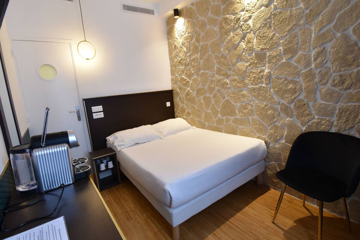 A hotel room with a bed, a chair, a coffee machine, a small table, and a stone wall.