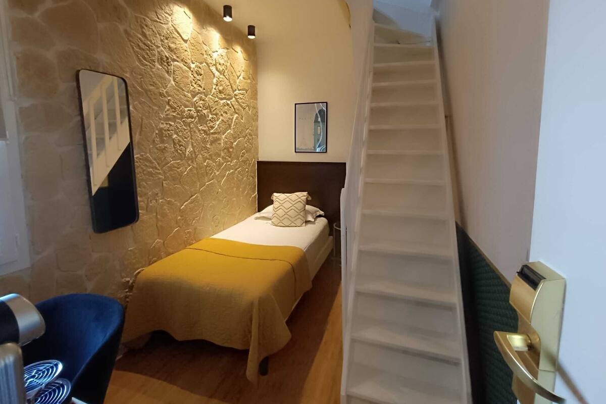 A bedroom with a stone wall, a bed with a yellow bedspread, a small desk with a chair, and a staircase.
