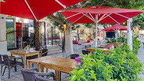 Un café al aire libre con sombrillas rojas, mobiliario de mimbre y piso de baldosas.