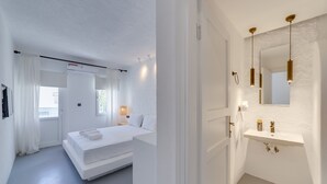 Un dormitorio moderno con una cama grande, una mesita de noche y una ventana con cortinas. Hay un baño con lavamanos, espejo y luces colgantes.