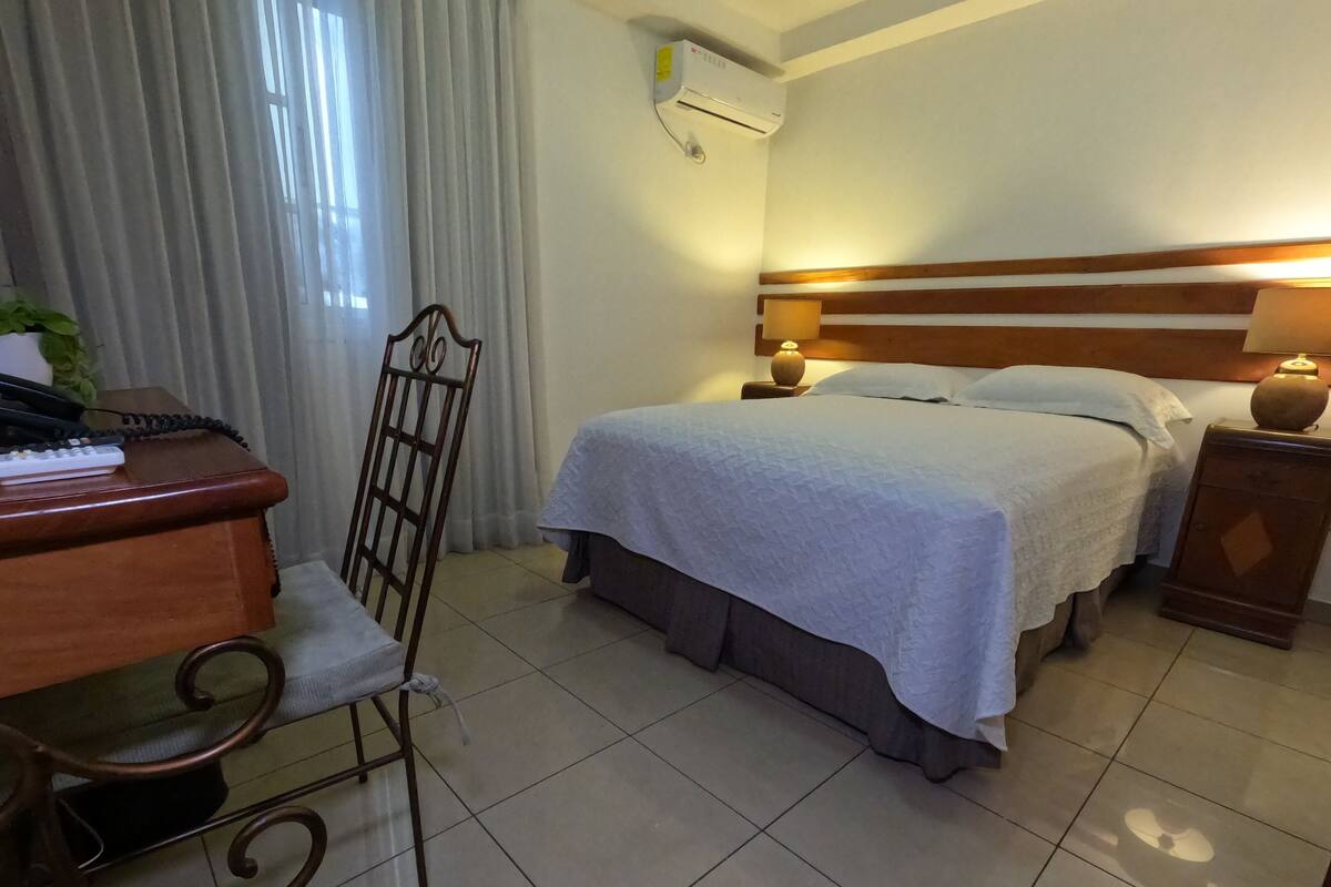 Habitación de hotel con cama, escritorio con teléfono, silla, mesita de noche con lámpara y unidad de aire acondicionado en la pared.