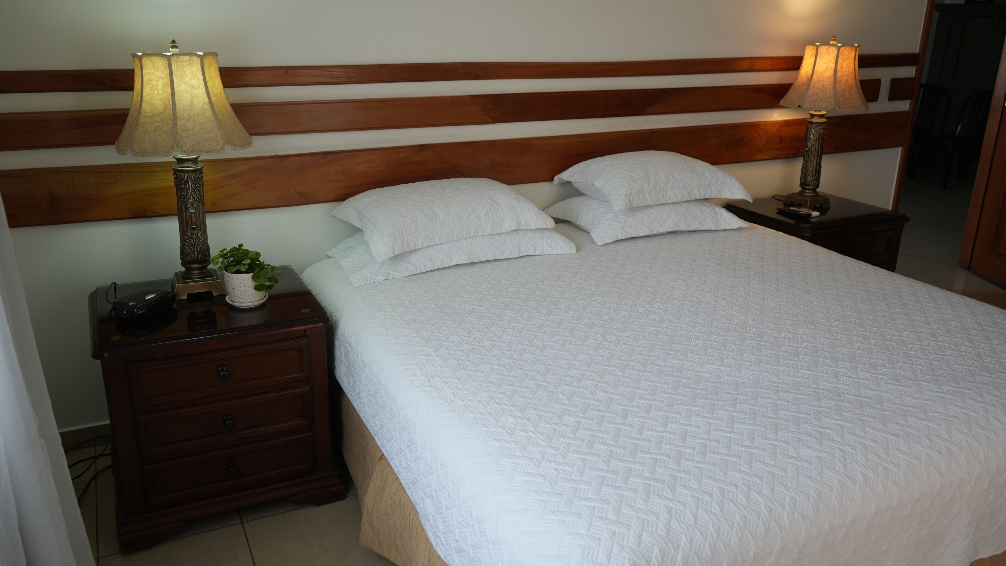 A neatly made bed with white linens, two bedside lamps, a nightstand with a plant, and a wooden headboard.