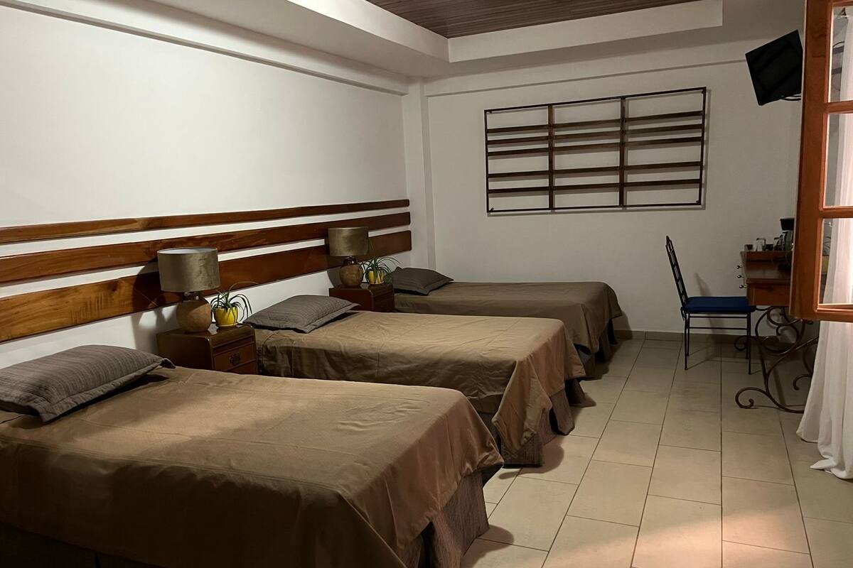 Habitación de hotel con tres camas individuales, un escritorio con silla, un televisor fijado a la pared y una ventana con persianas.