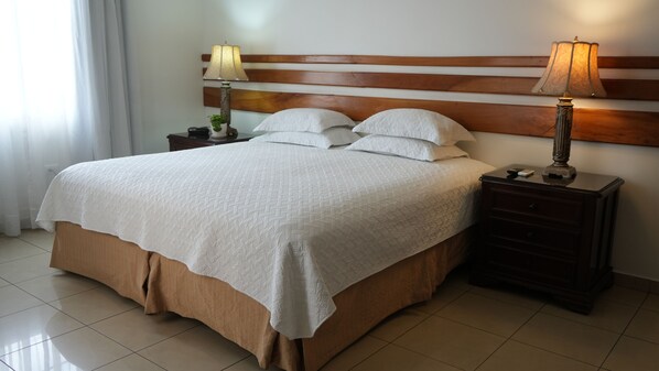 Una cama bien hecha con cabecera de madera, dos mesitas de noche con lámparas y suelo de baldosas.