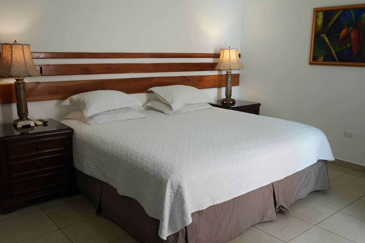 Una cama bien hecha con cabecera de madera, dos mesitas de noche con lámparas y un cuadro en la pared.