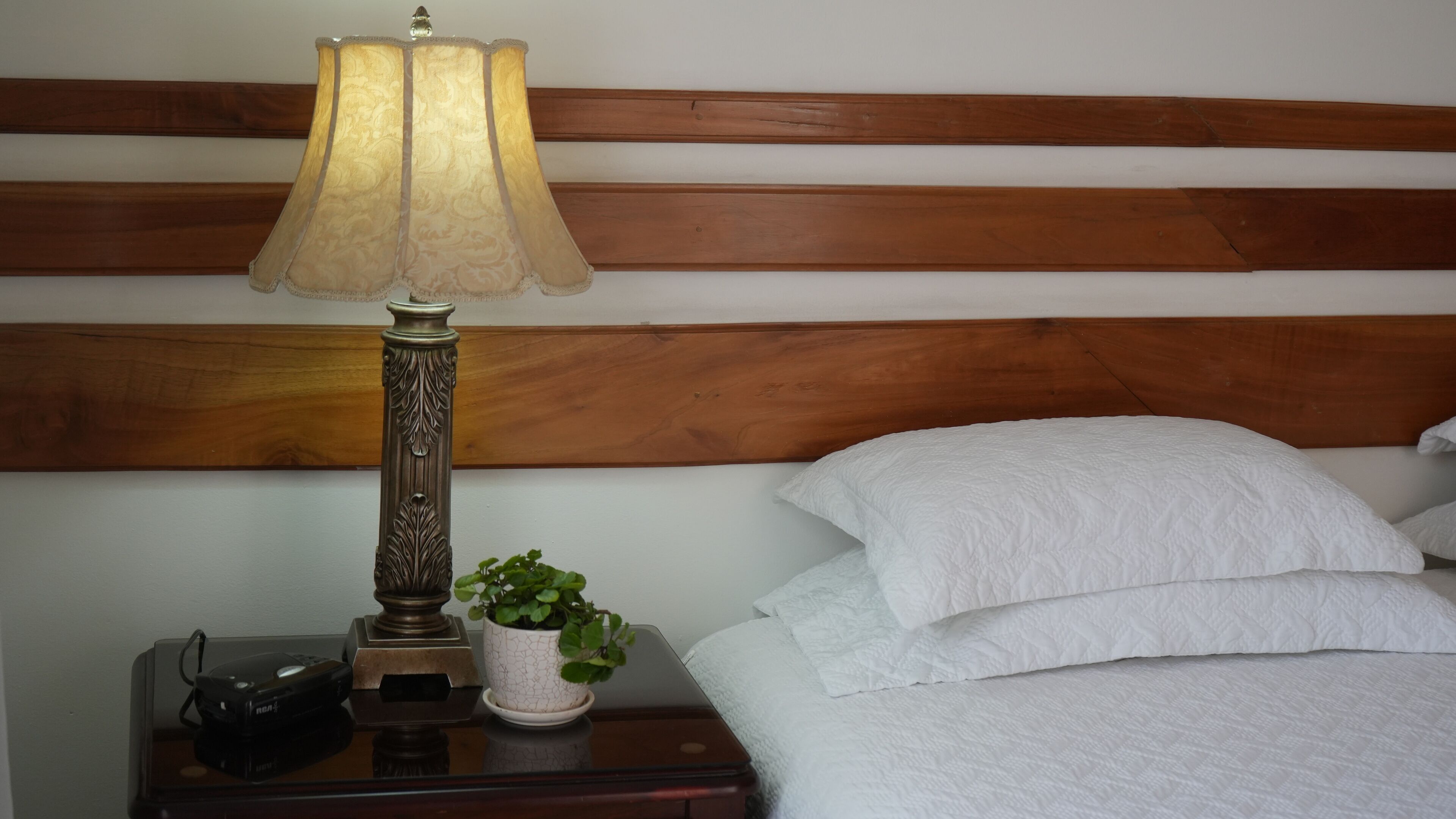 Una cama bien hecha con sábanas blancas, un cabecero de madera, una mesita de noche con una lámpara, una pequeña planta en maceta y un control remoto.