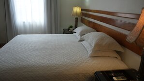 Uma cama bem arrumada com um edredom branco acolchoado, uma cabeceira de madeira, uma luminária de mesa e uma pequena mesa com um controle remoto.