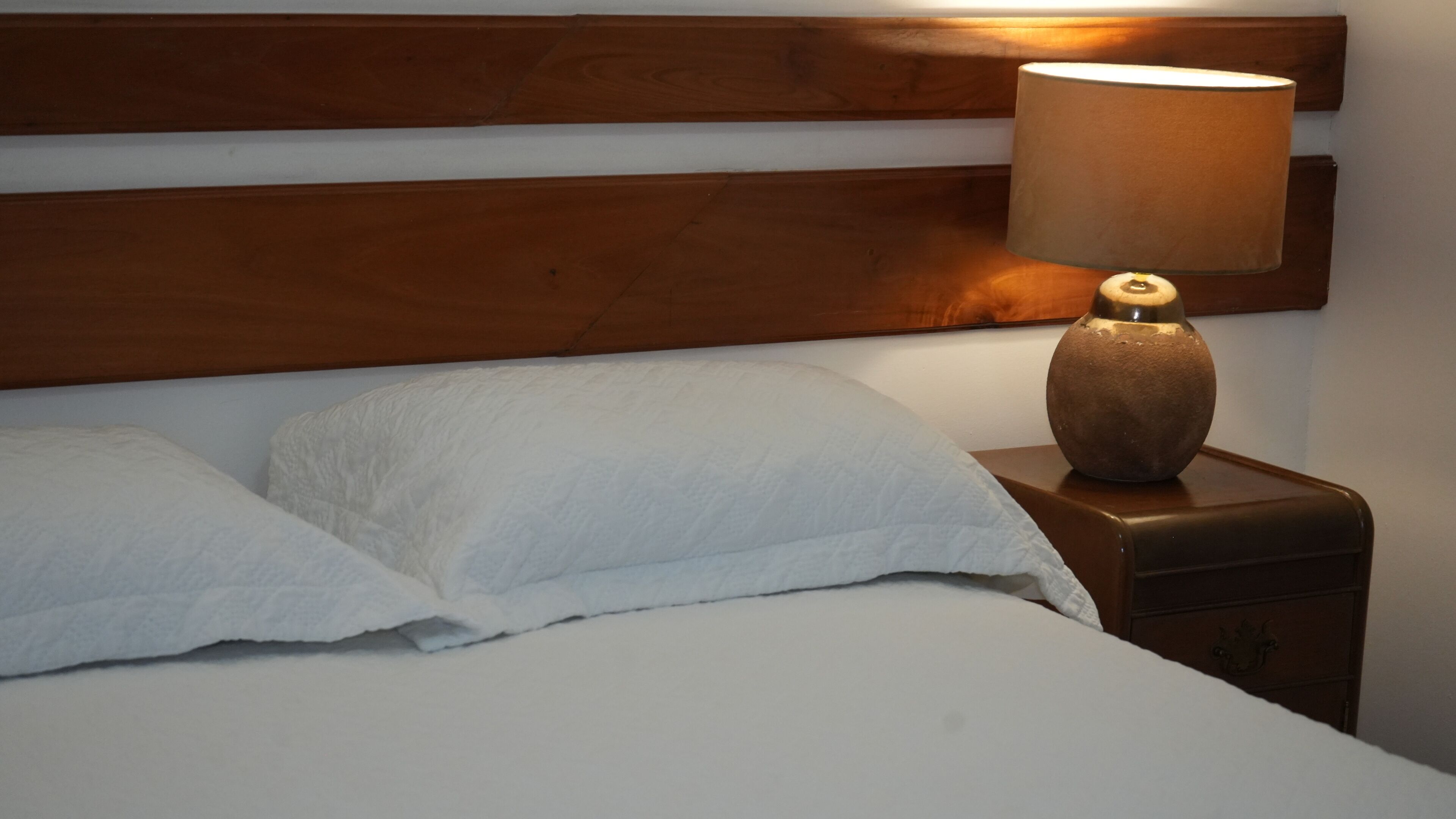 A bed with white linens, a wooden headboard, and a bedside lamp on a wooden nightstand.
