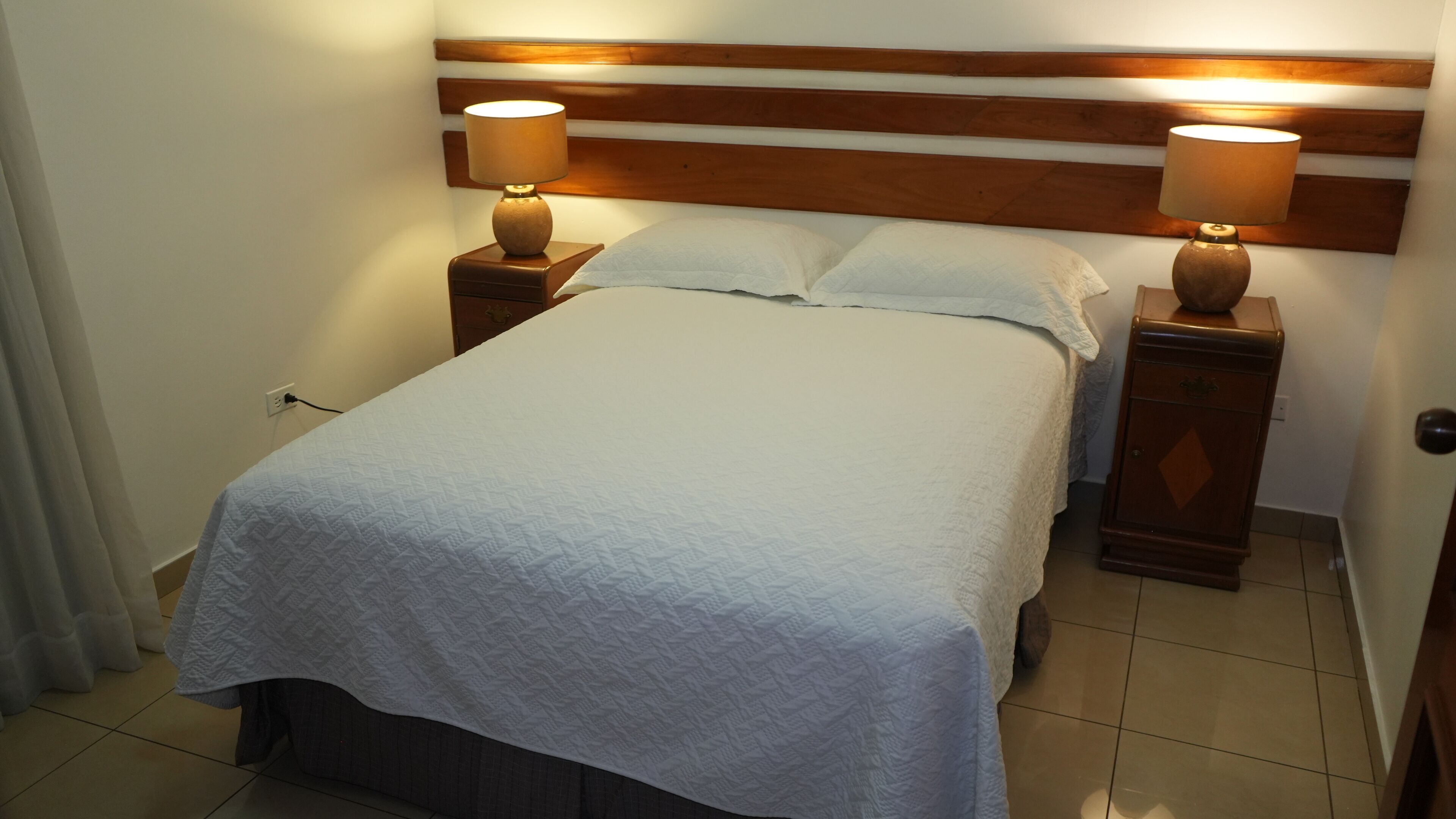 A neatly made bed with a white quilt, flanked by two bedside tables with lamps, in a room with a wooden headboard and tiled floor.