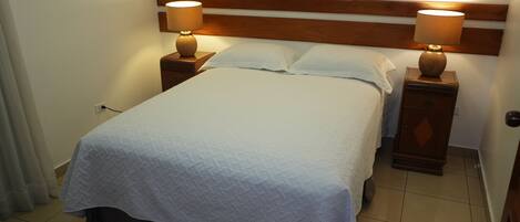 A neatly made bed with a white quilt, flanked by two bedside tables with lamps, in a room with a wooden headboard and tiled floor.