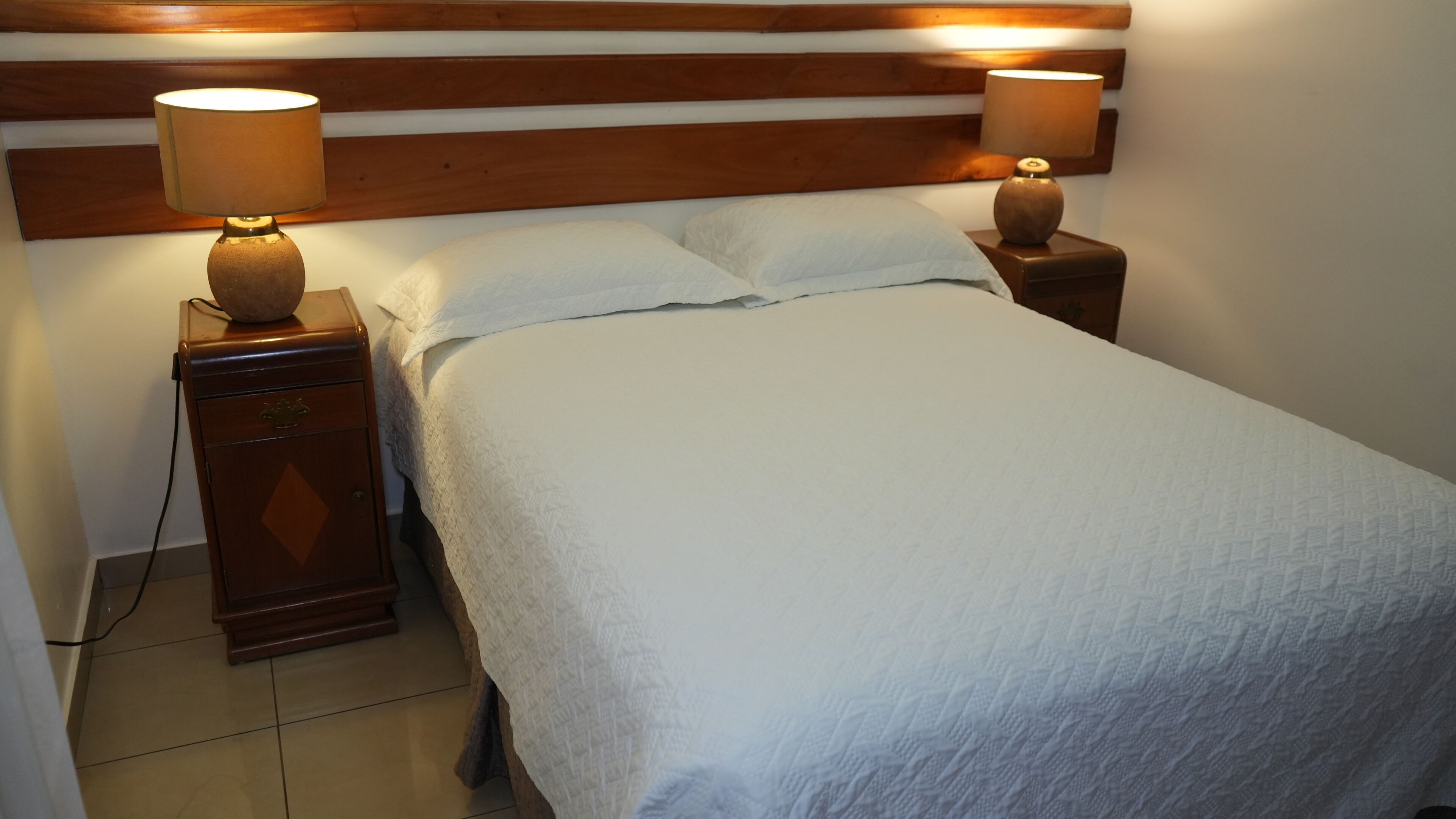 Una cama bien hecha con cabecera de madera, dos lámparas de noche y una mesita de noche de madera con una lámpara.