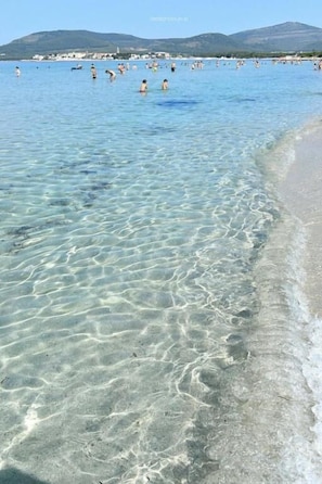 Una platja amb aigües poc profundes i cristal·lines, i una cadena de muntanyes a la distància.