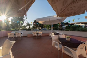 Terrassa exterior amb caires i taules de plàstic blanc, coberta per grans paraigües, i un cel blau al fons.