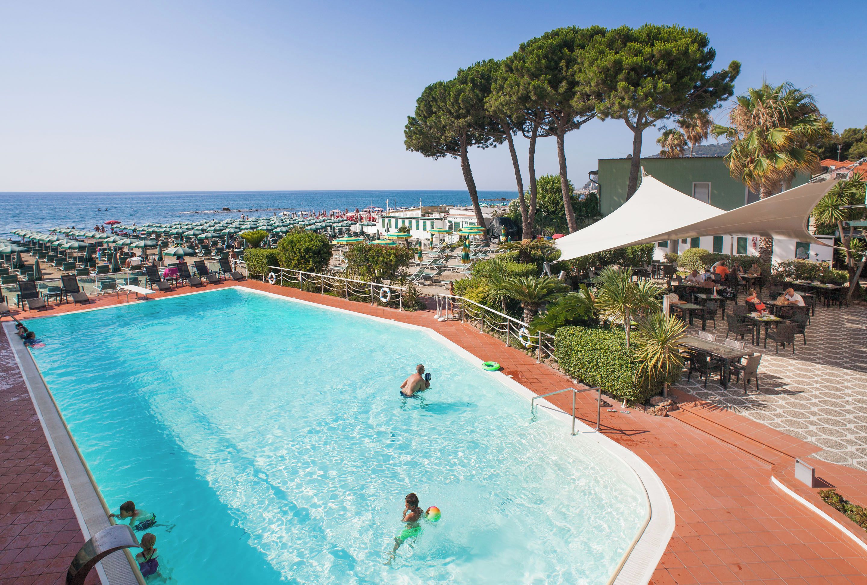 A swimming pool with a view of the sea, surrounded by beach umbrellas and lounge chairs.
