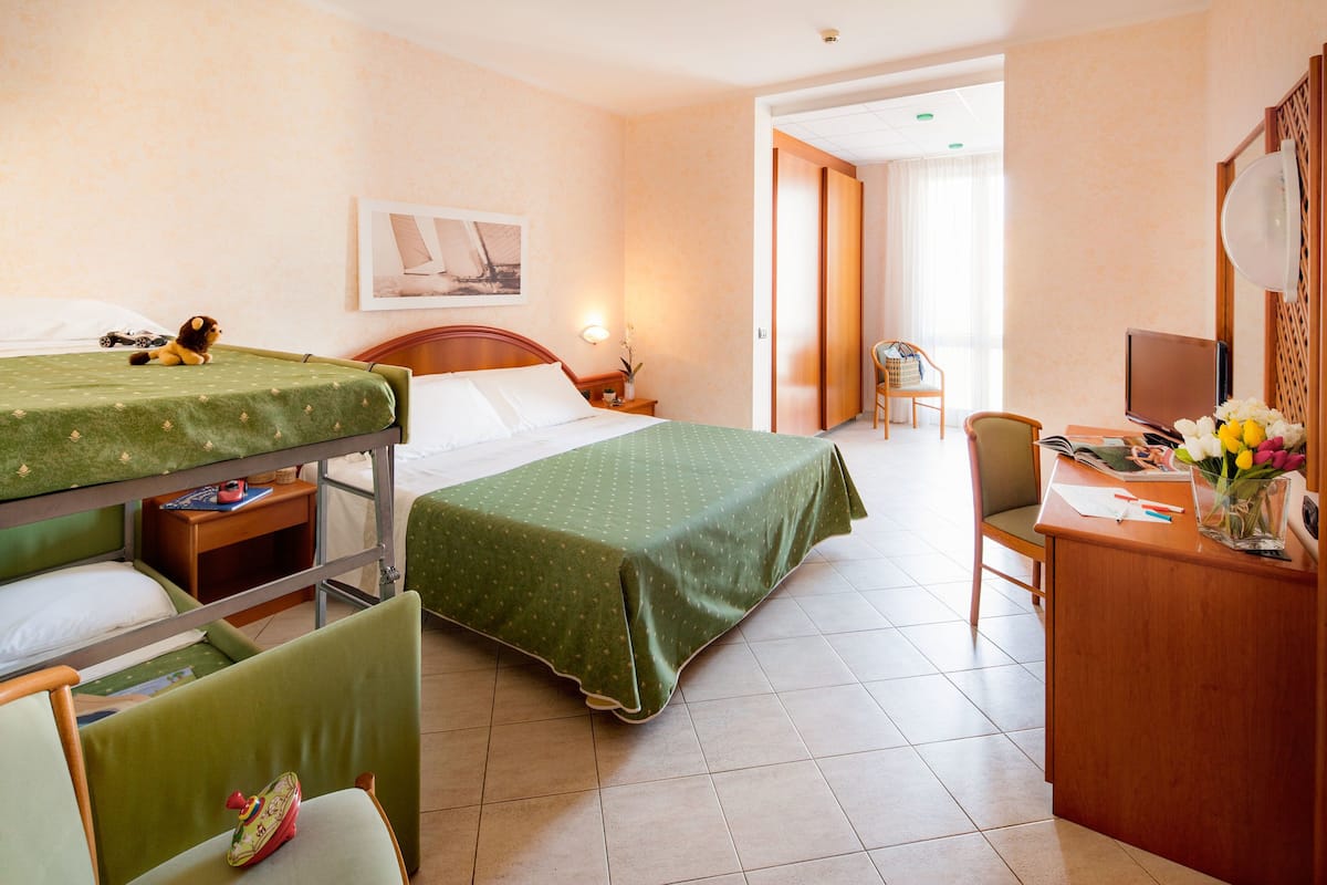 A hotel room with a single bed, a desk with a computer, a chair, and a small table with a lamp.
