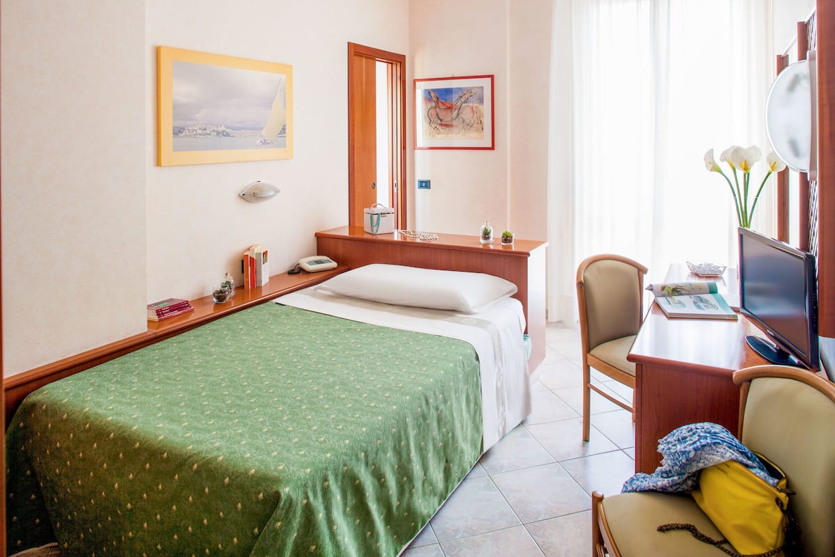 A hotel room with a bed, a desk with a computer and a chair, a bedside table with a lamp, and a window with curtains.