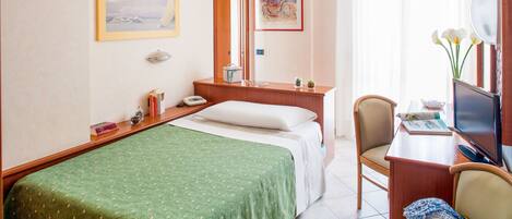 A hotel room with a bed, a desk with a computer and a chair, a bedside table with a lamp, and a window with curtains.
