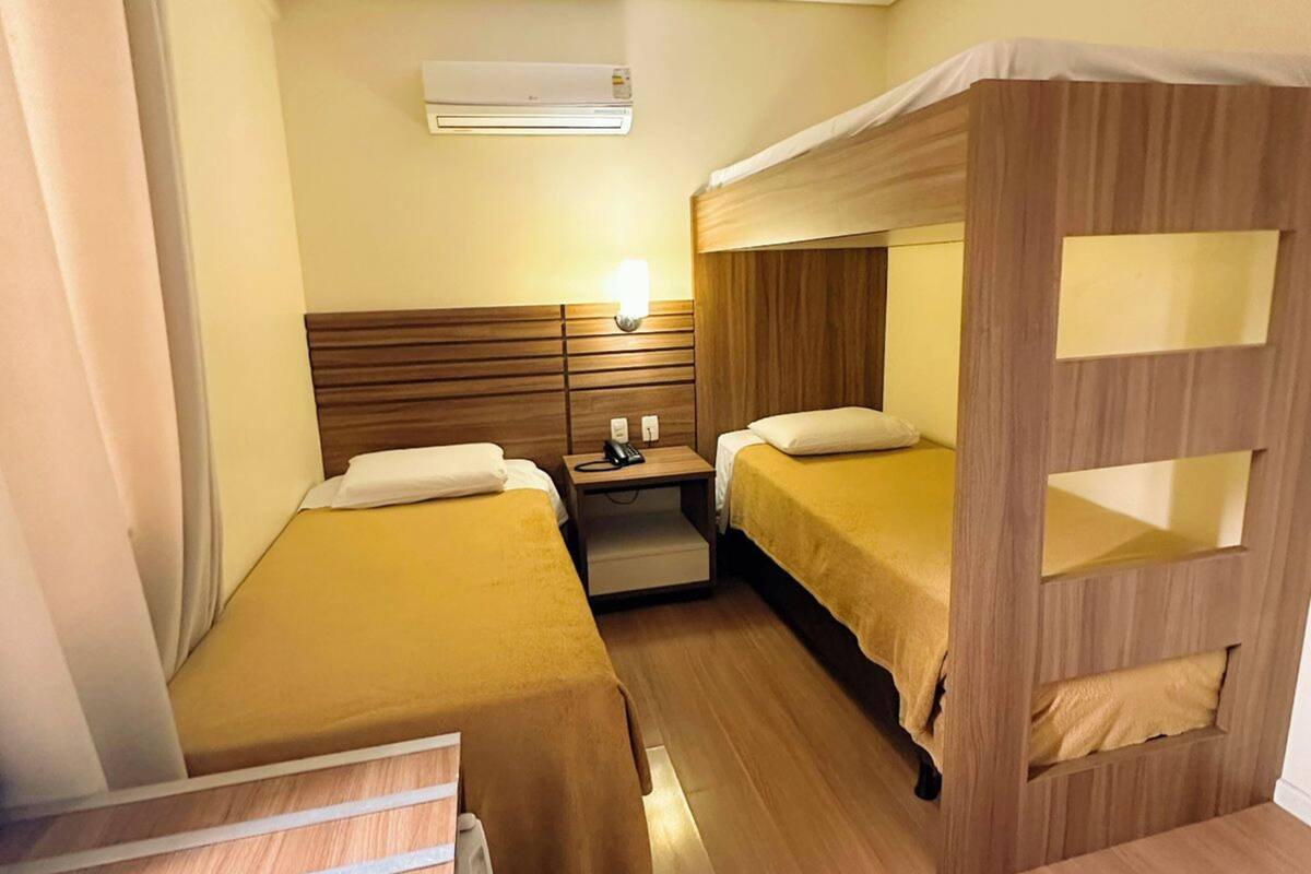 A compact hotel room with two single beds, a wooden bunk bed, a small bedside table with a lamp, and an air conditioning unit on the wall.
