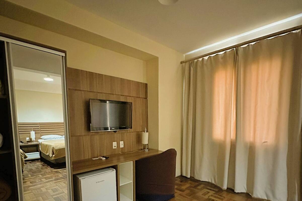 A hotel room with a wooden floor, a TV mounted on a wooden panel, a small fridge, a chair, and a bed in the background.
