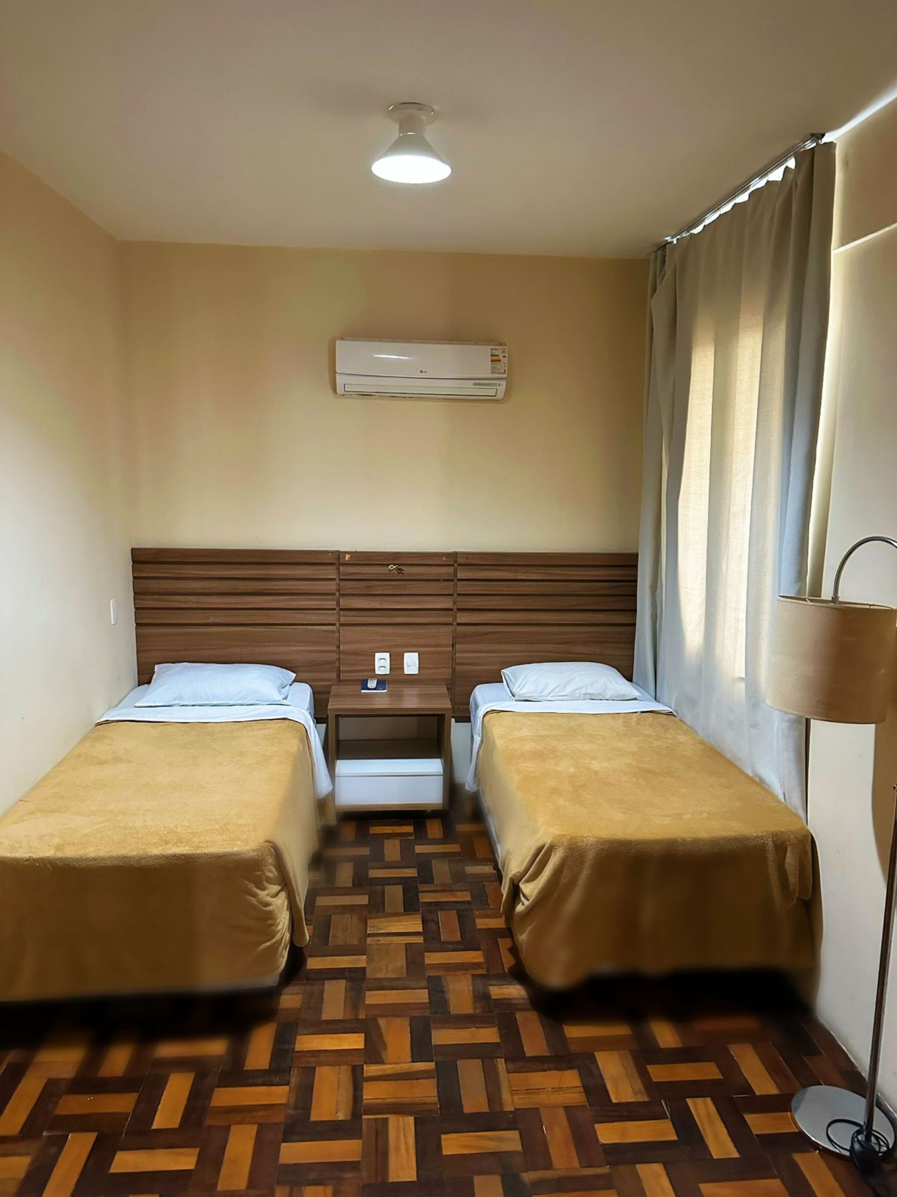 A hotel room with two single beds, a wooden headboard, an air conditioning unit, a bedside table with a phone, a patterned floor, and a floor lamp.