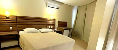 A hotel room with a large bed, bedside tables, a nightstand, an air conditioning unit, and a curtain on the window.