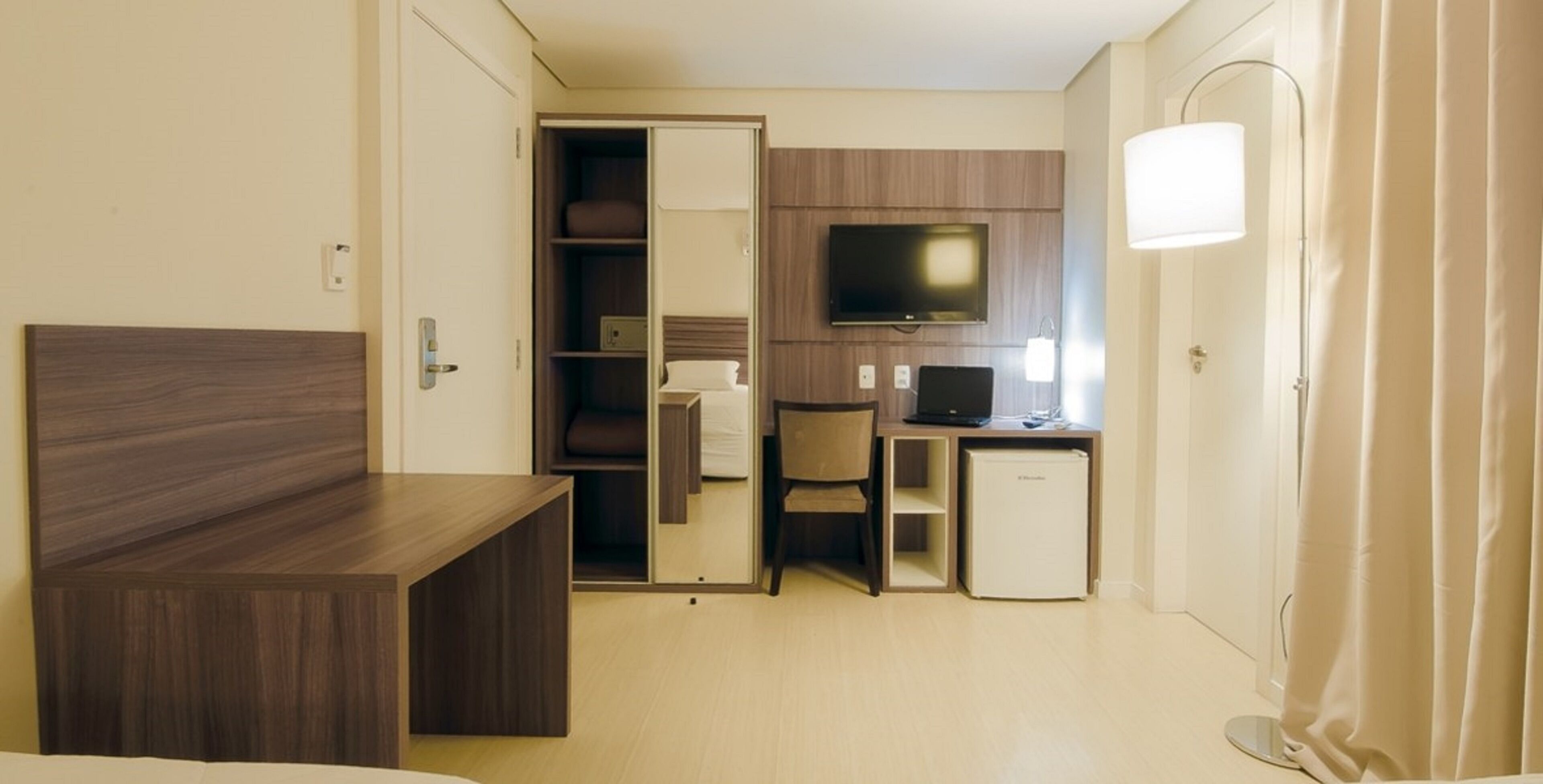 A compact hotel room with a bed, a desk, a television, a mini-fridge, and a wardrobe.