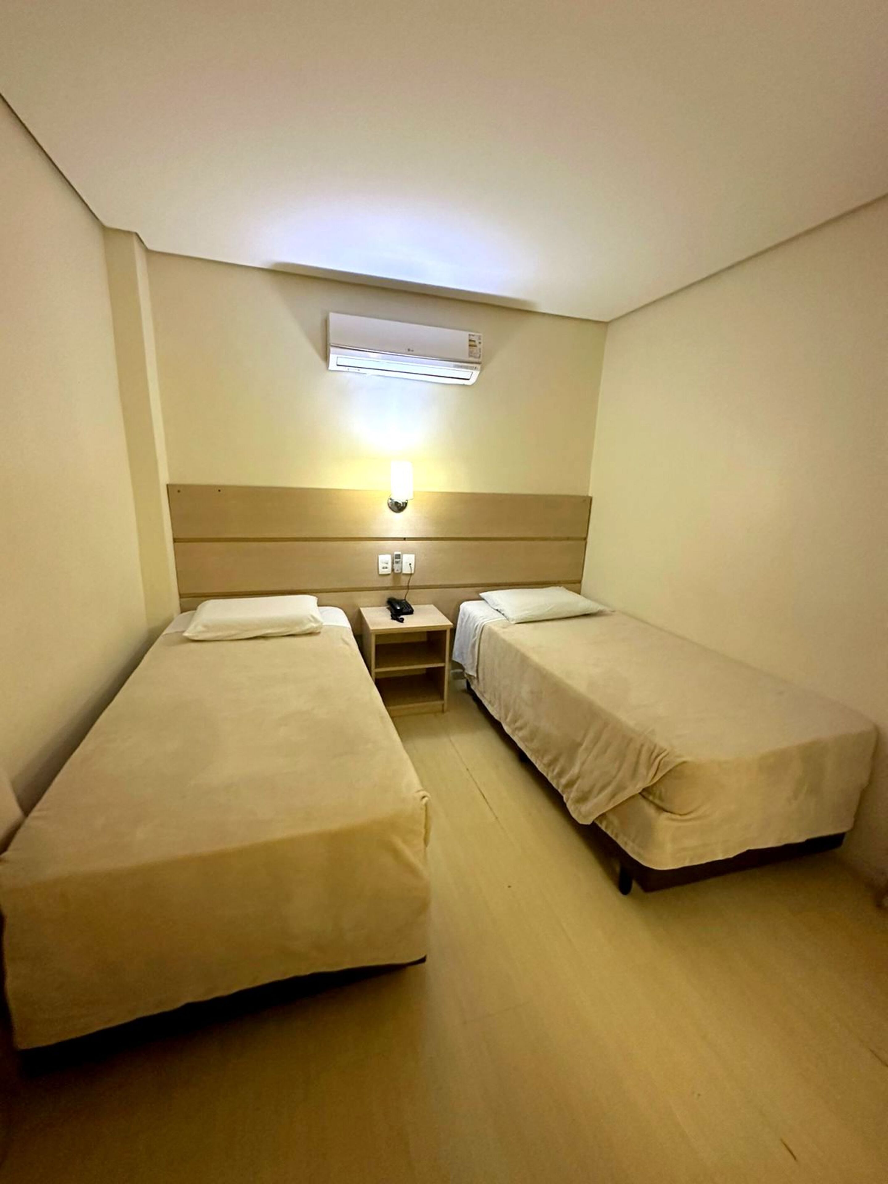 Une petite chambre d’hôtel comprenant une seule pièce, avec deux lits simples, une table de chevet avec une lampe, un climatiseur fixé au mur et une prise de courant.