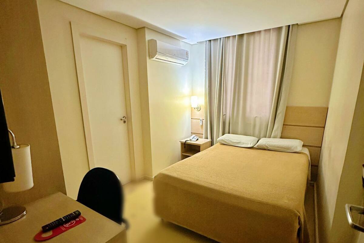 A hotel room with a bed, a desk with a lamp, a chair, an air conditioning unit, and a nightstand with a phone.