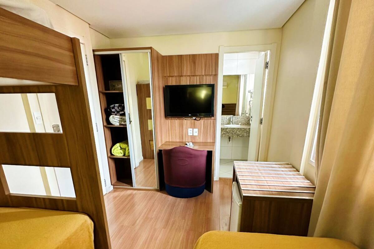 A compact hotel room with wooden flooring, a television mounted on the wall, a small kitchenette, and a bed with a yellow cover.