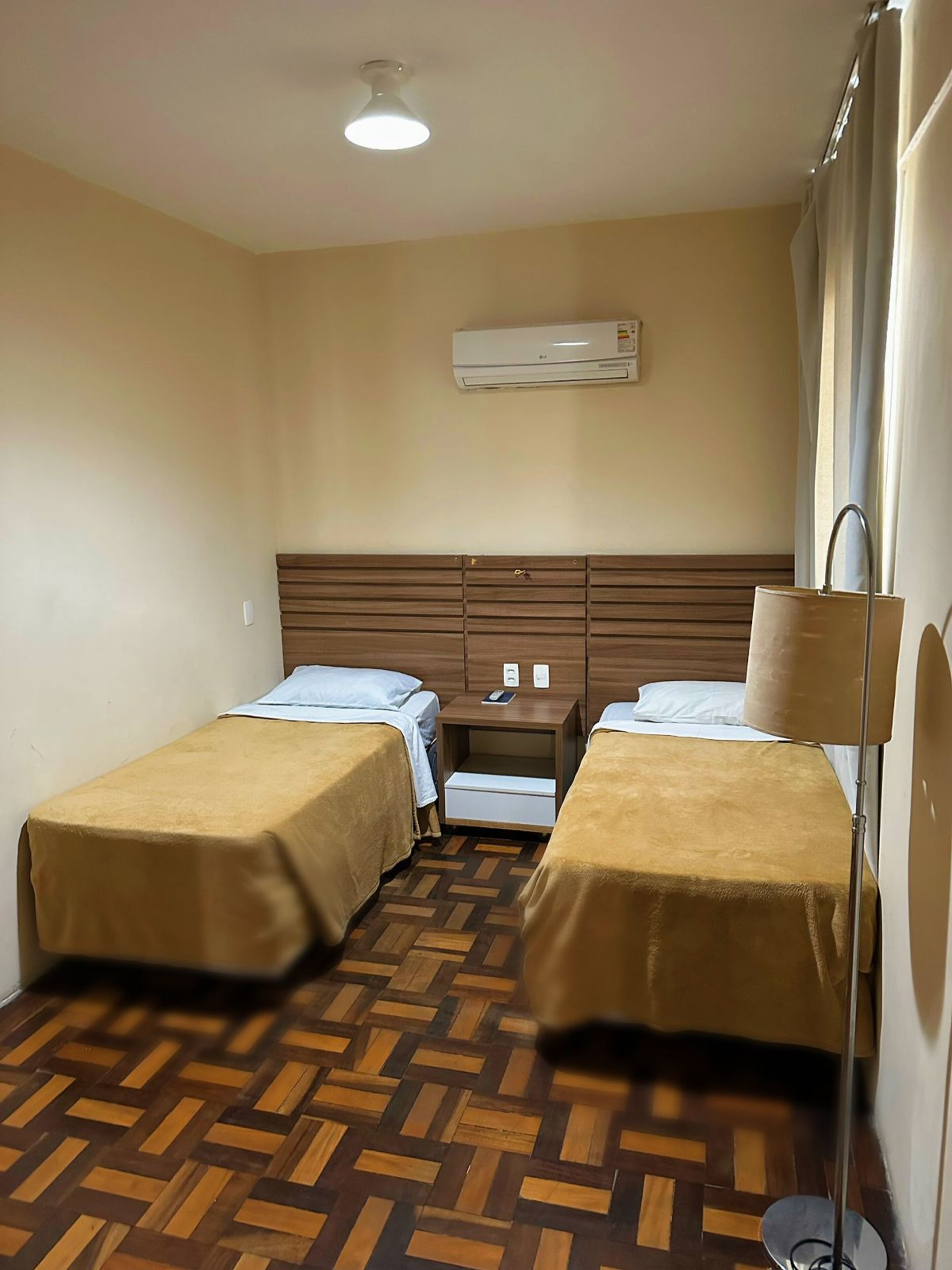 A hotel room with two beds, a wooden floor, an air conditioning unit, a nightstand, and a lamp.