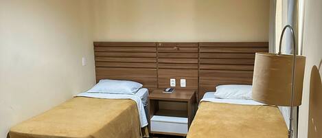 A hotel room with two beds, a wooden floor, an air conditioning unit, a nightstand, and a lamp.