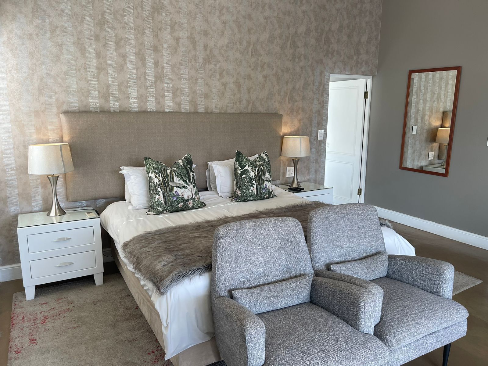 A bedroom with a large bed, two nightstands with lamps, a grey armchair, a fur throw, and a mirror on the wall.