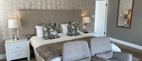 A bedroom with a large bed, two nightstands with lamps, a grey armchair, a fur throw, and a mirror on the wall.