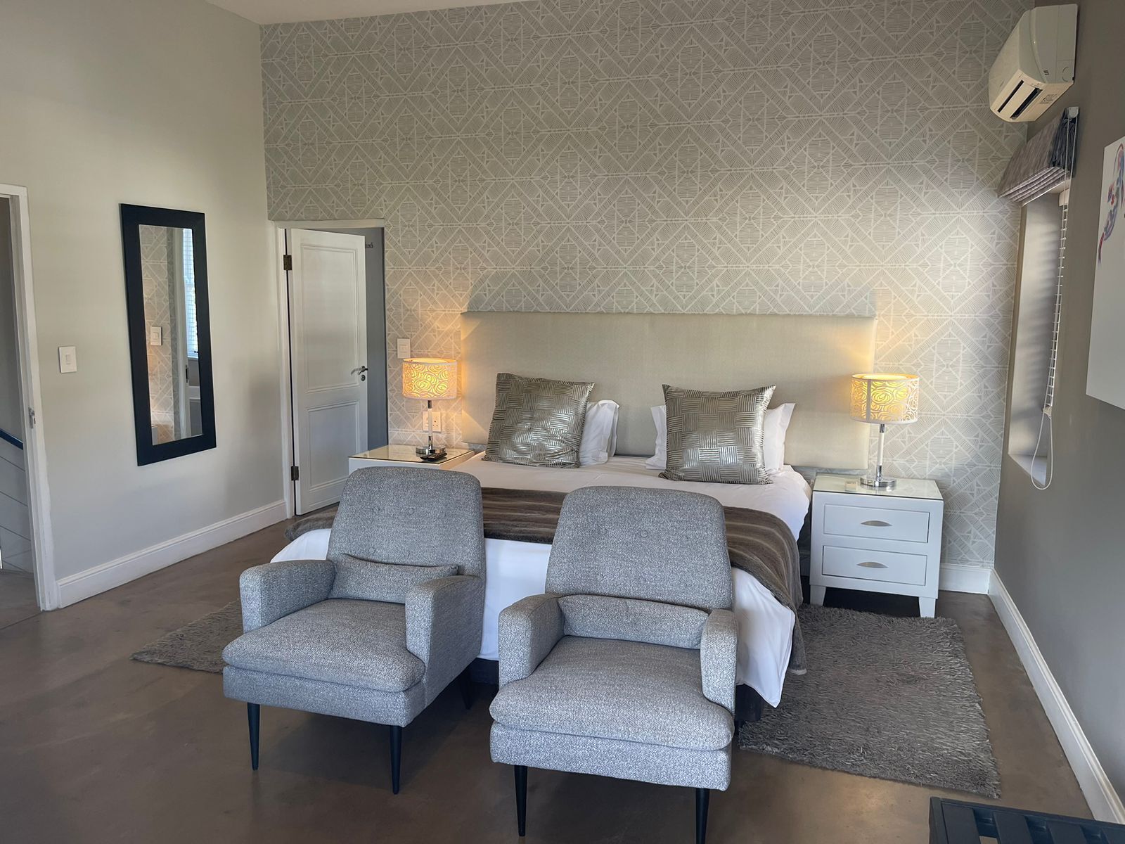 A hotel room with a bed, two armchairs, a nightstand, a mirror, and patterned wallpaper.