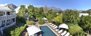 Zona de piscina con sillones de descanso y sombrillas, rodeada de exuberante vegetación y montañas al fondo.