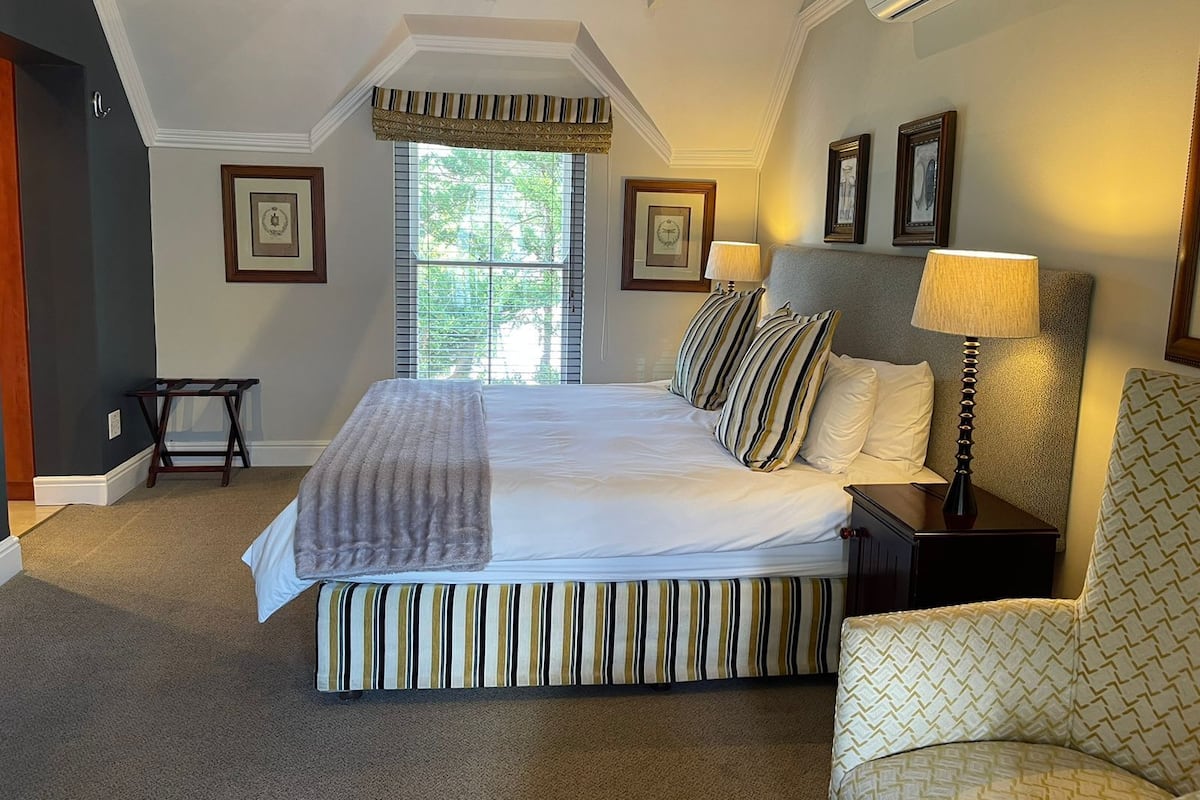 A bedroom with a large bed, a chair, a nightstand, a window with blinds, and framed pictures on the wall.
