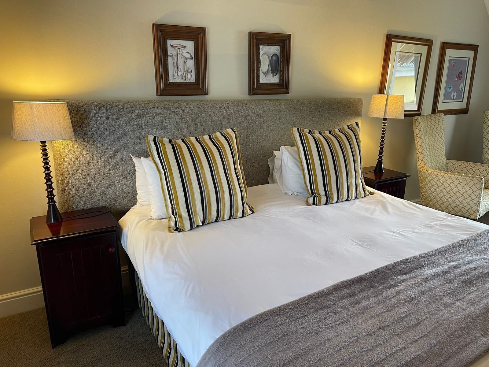 Een hotelkamer met een groot bed, twee nachtkastjes en een stoel. Boven het bed hangen afbeeldingen in lijsten.