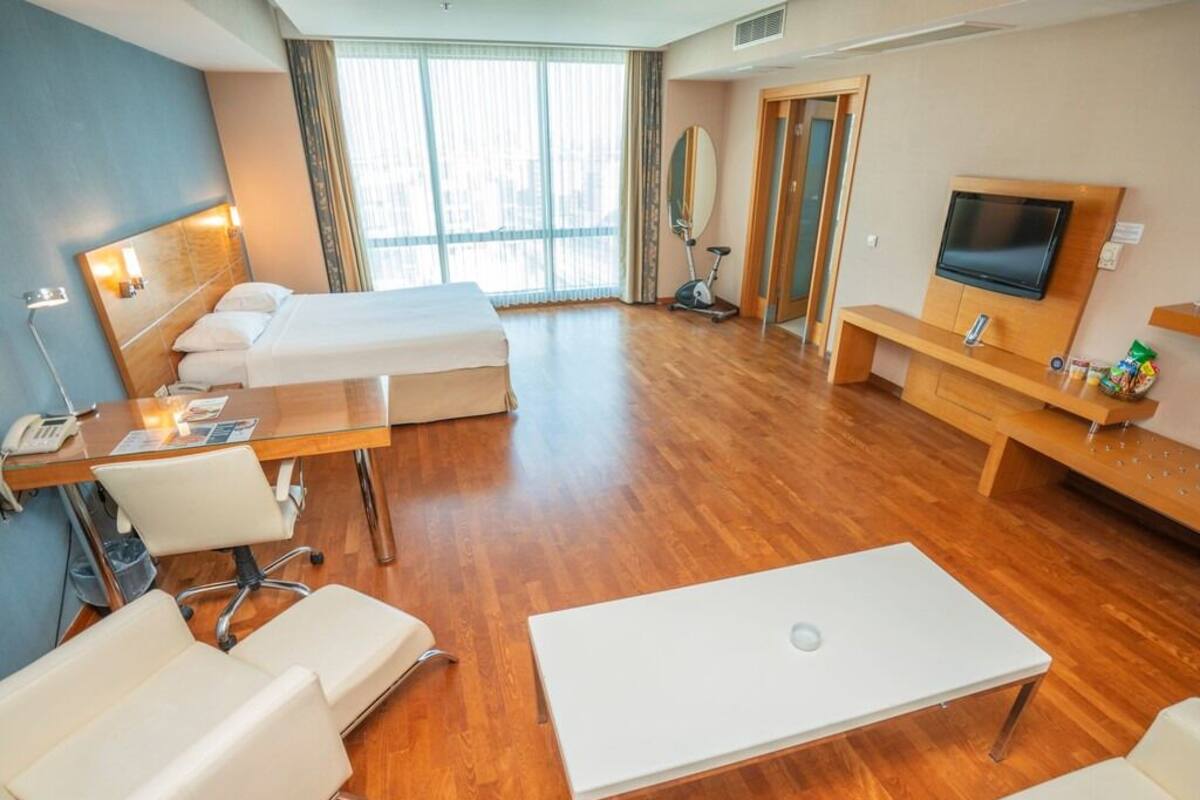 A hotel room with a bed, two white sofas, a desk with a lamp, a TV, and a treadmill.