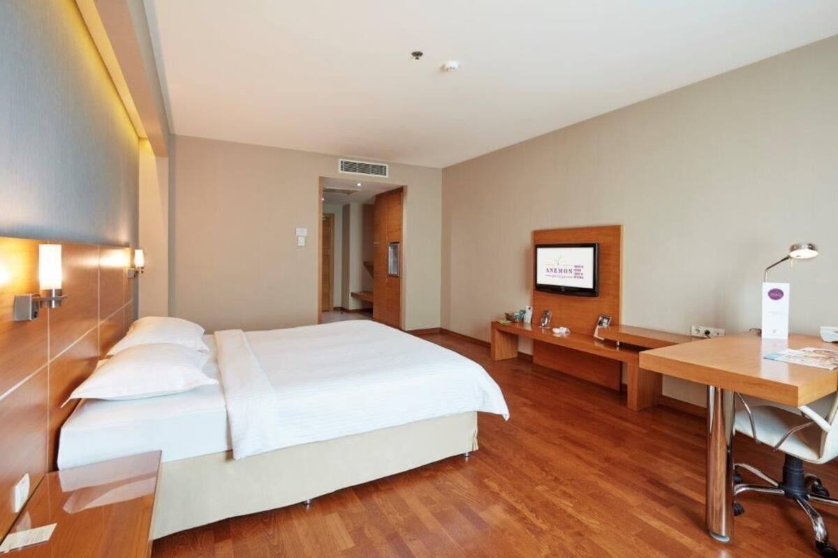 A hotel room with a bed, a desk, a chair, a lamp, a TV, and a small table with a vase.