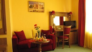 A hotel room with a red sofa, a small wooden table with a fruit platter, a TV, and a desk with a chair. - Hotel am Hauptbahnhof (Schwerin)
