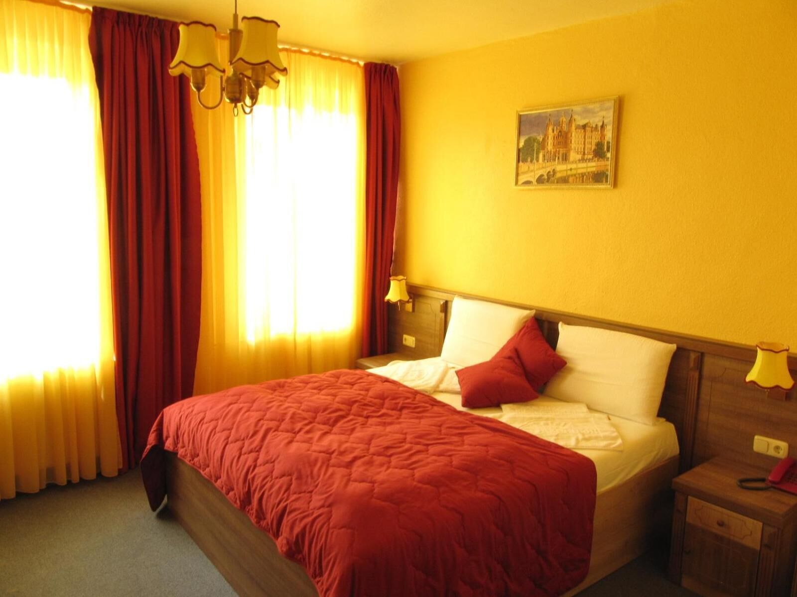 A hotel room with a large bed, red curtains, a painting on the wall, and a nightstand with a lamp.