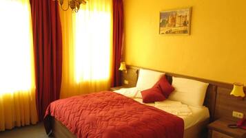 A hotel room with a large bed, red curtains, a painting on the wall, and a nightstand with a lamp.