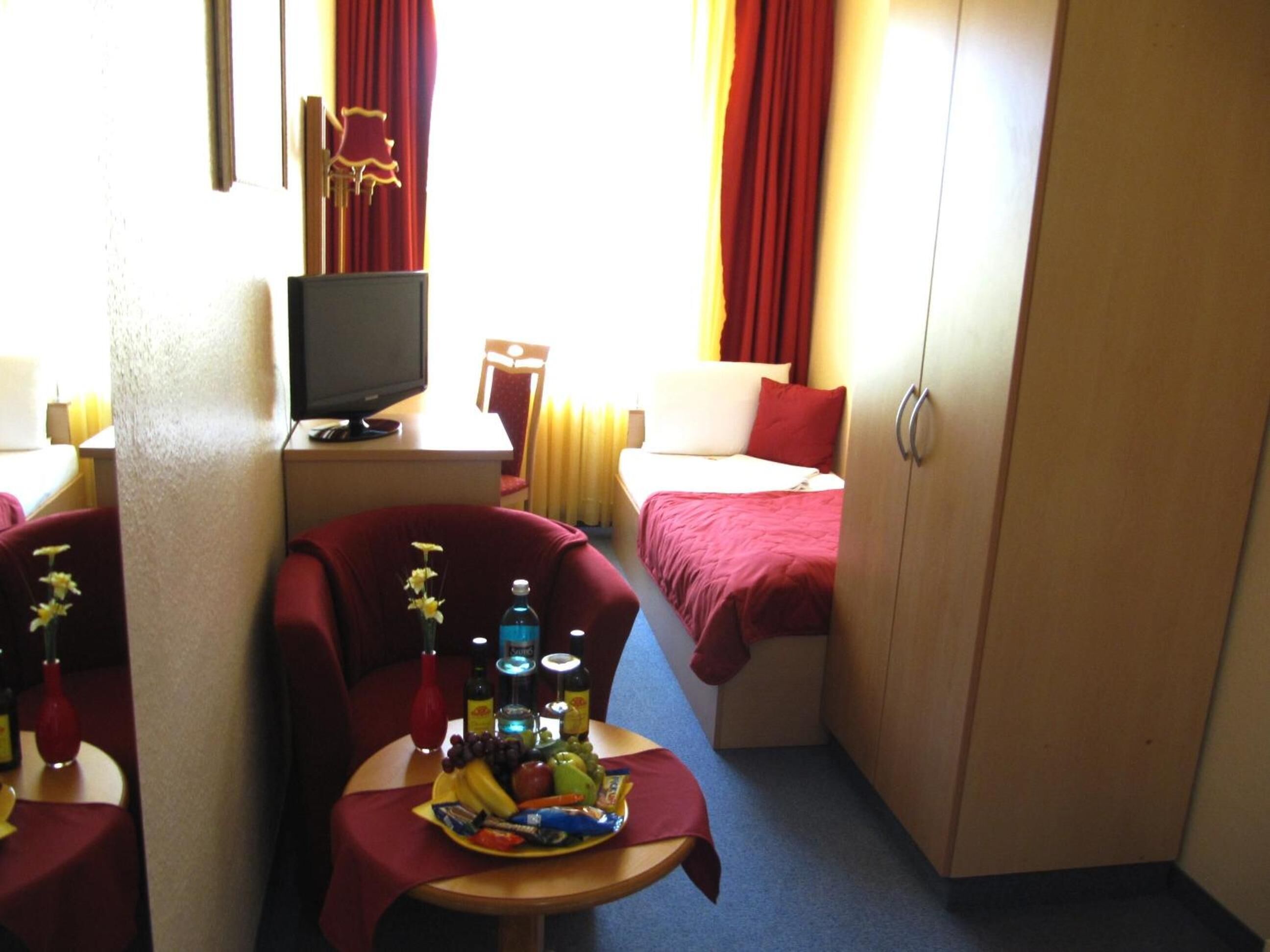 Una habitación de hotel funcional con una cama, un escritorio con televisor, una silla y una pequeña mesa con una bandeja de frutas.