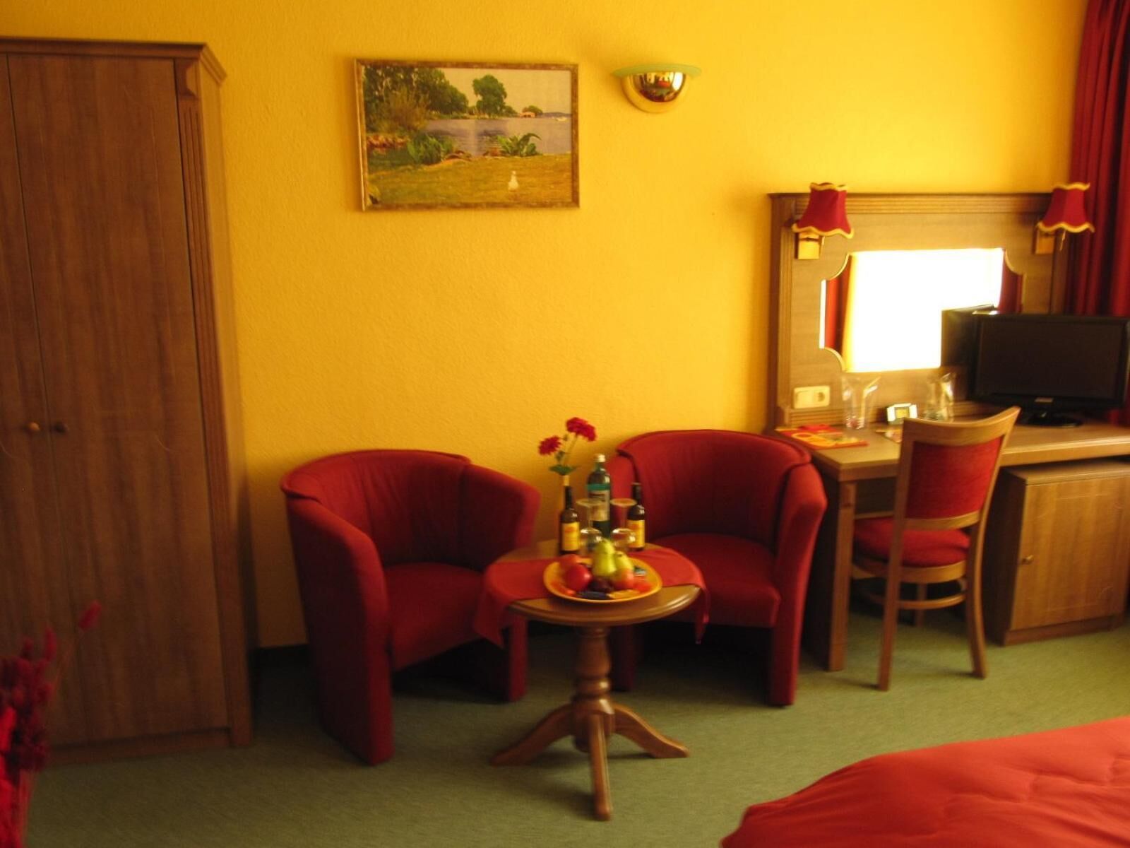 Habitació d'hotel amb un armari de fusta, dues butaques vermelles, una taula rodona petita amb un plat de fruita, un mirall, una televisió i una finestra amb cortines.