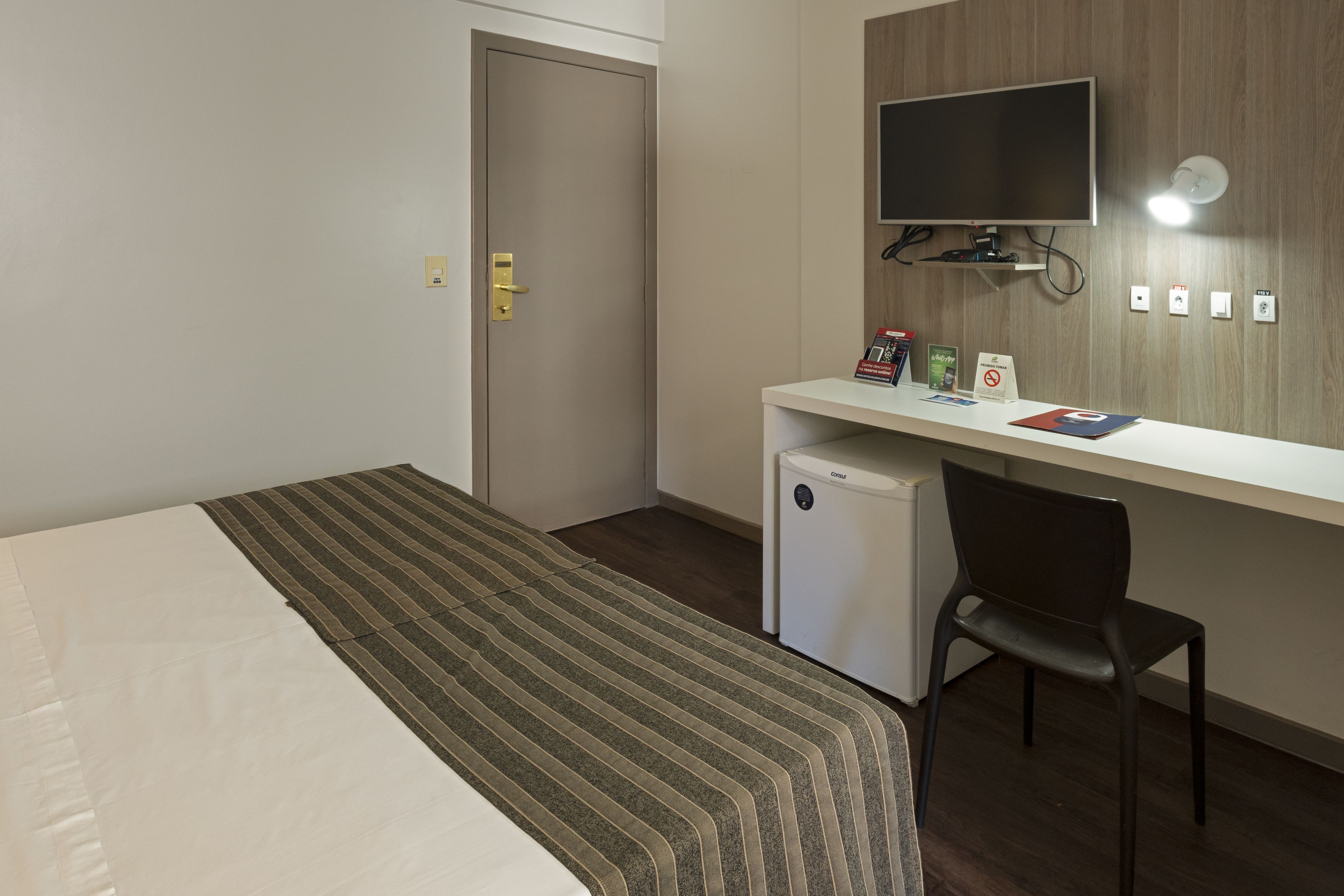 Une chambre d’hôtel équipée d’un lit, d’un bureau avec un ordinateur, d’une chaise, d’une télévision et d’un petit réfrigérateur.