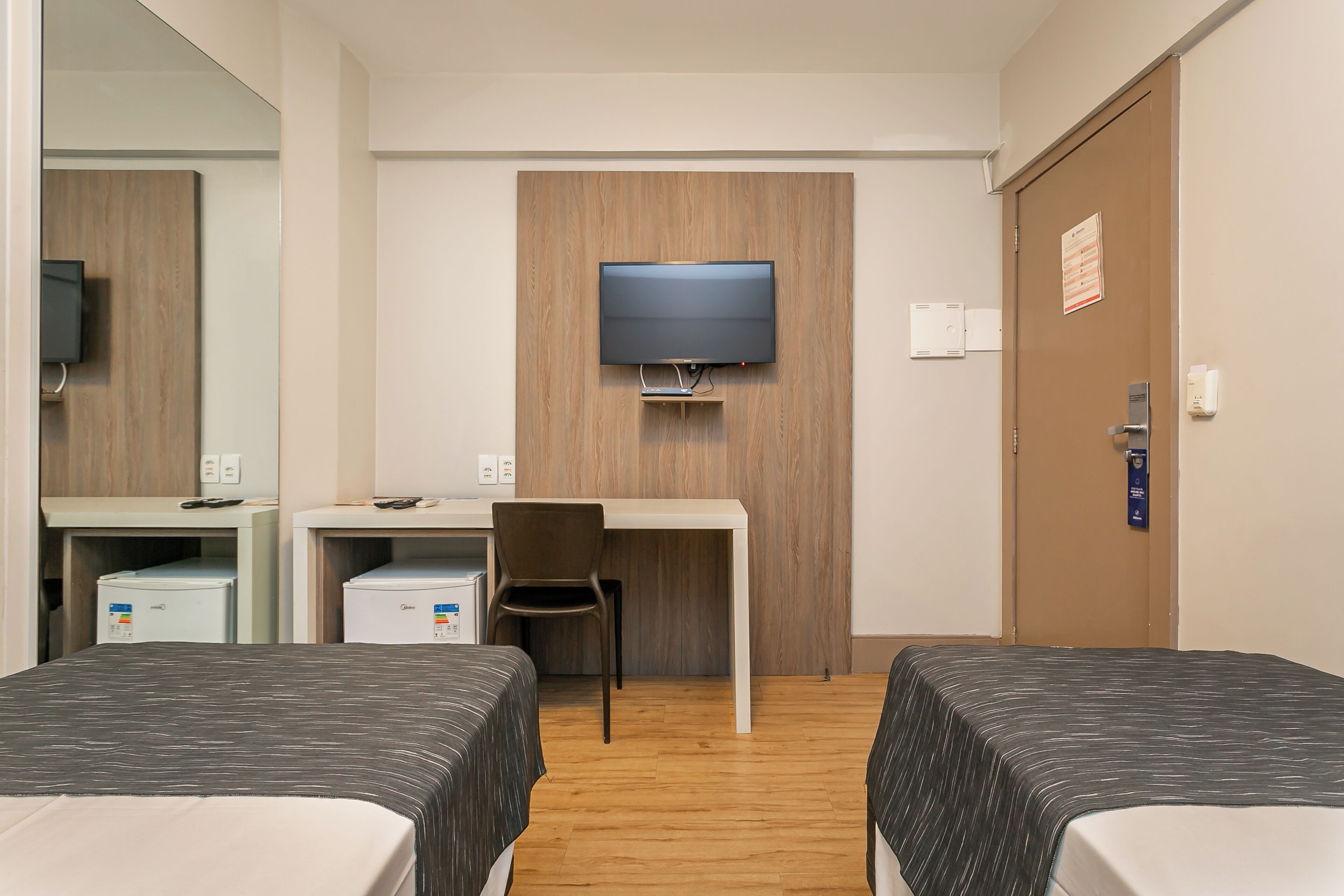 Ett hotellrum med två sängar, ett skrivbord, en stol, en tv som är monterad på väggen och en dörr med en skylt.