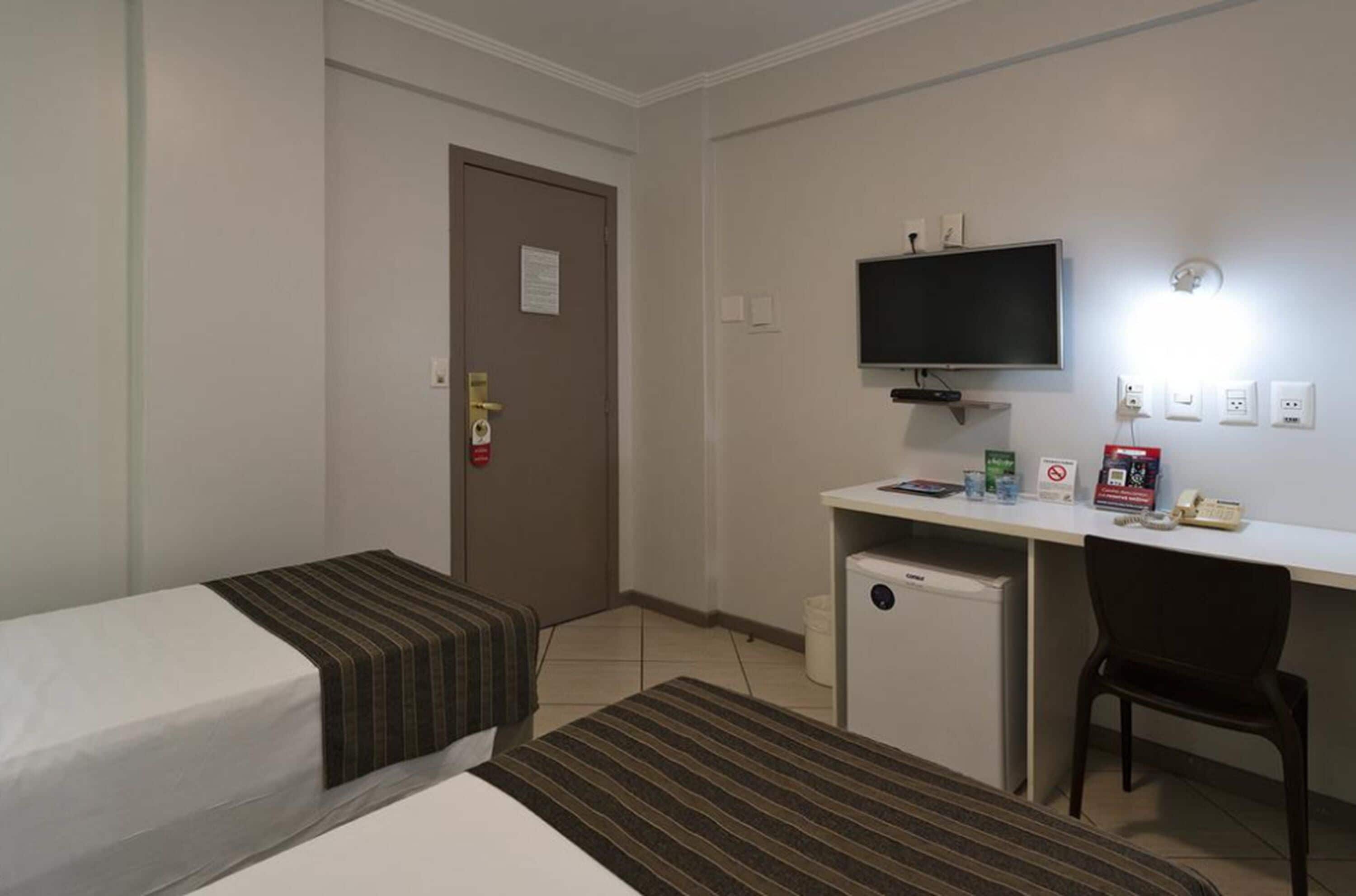 Quarto de hotel com duas camas de solteiro, uma escrivaninha com computador, televisão e um pequeno frigorífico.