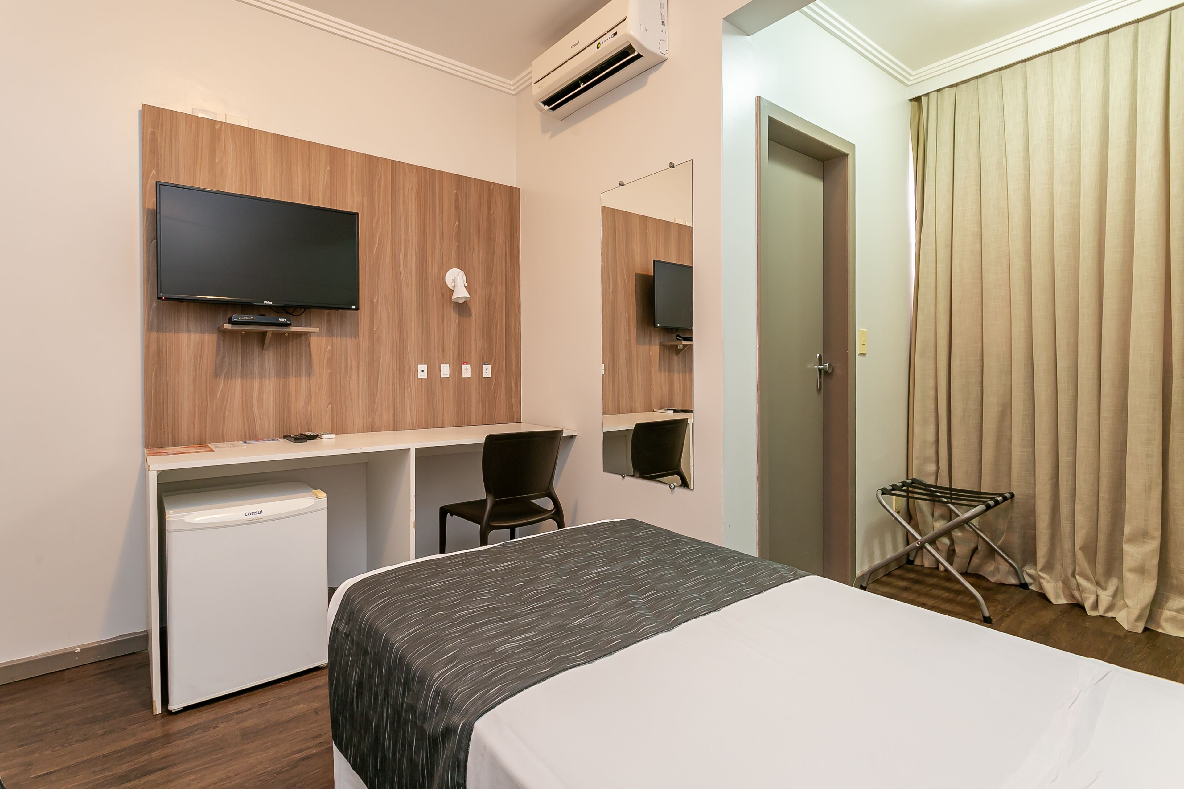 A hotel room with a bed, a desk with a television, a small refrigerator, a chair, and a mirror.